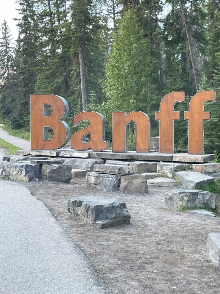 Brown B A N F F letters in large sign on stone wall against pine trees