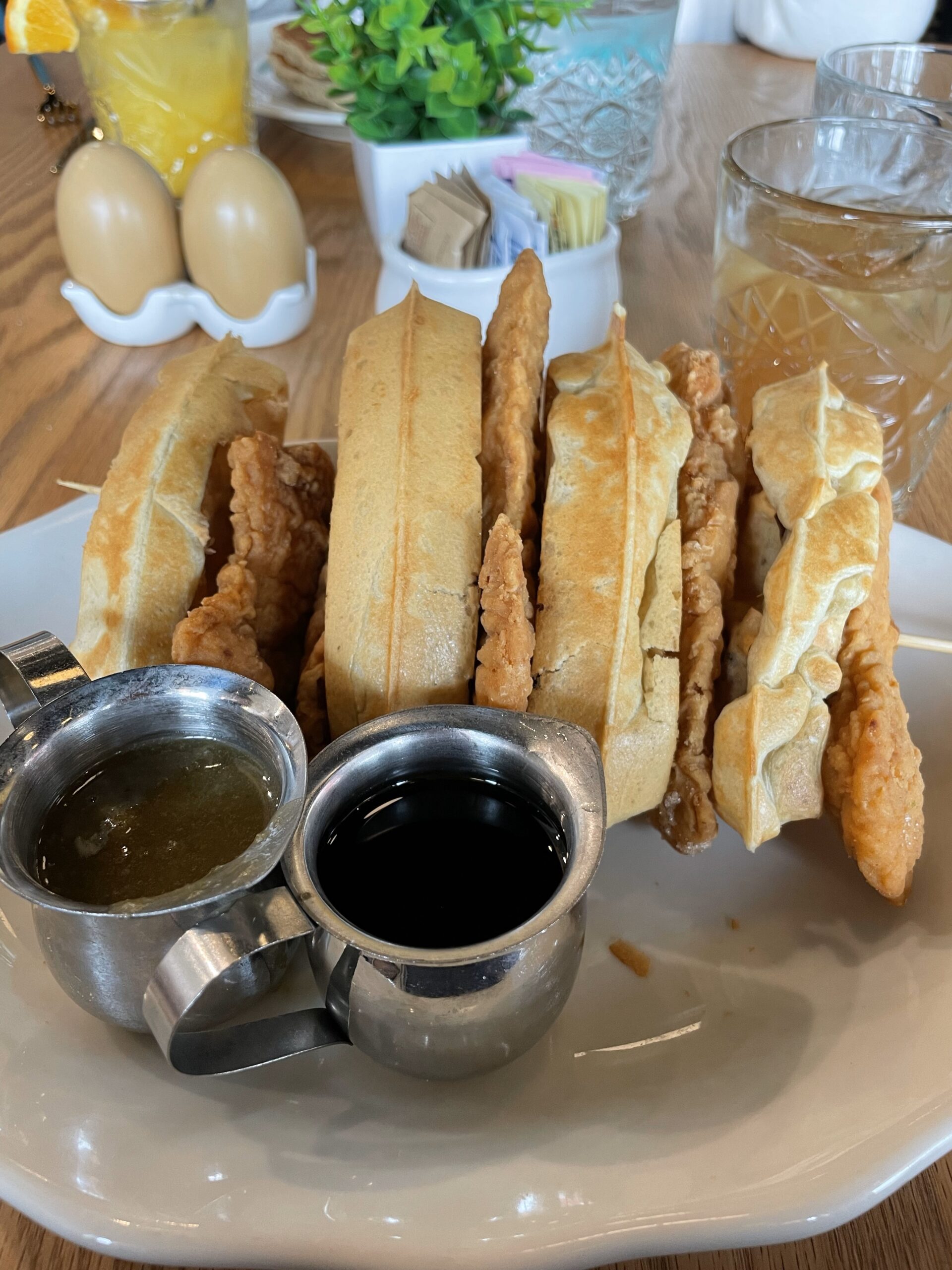 Waffles and Fried Chickens layered on a skewer with syrup and green sauce in silver cups on a white plate