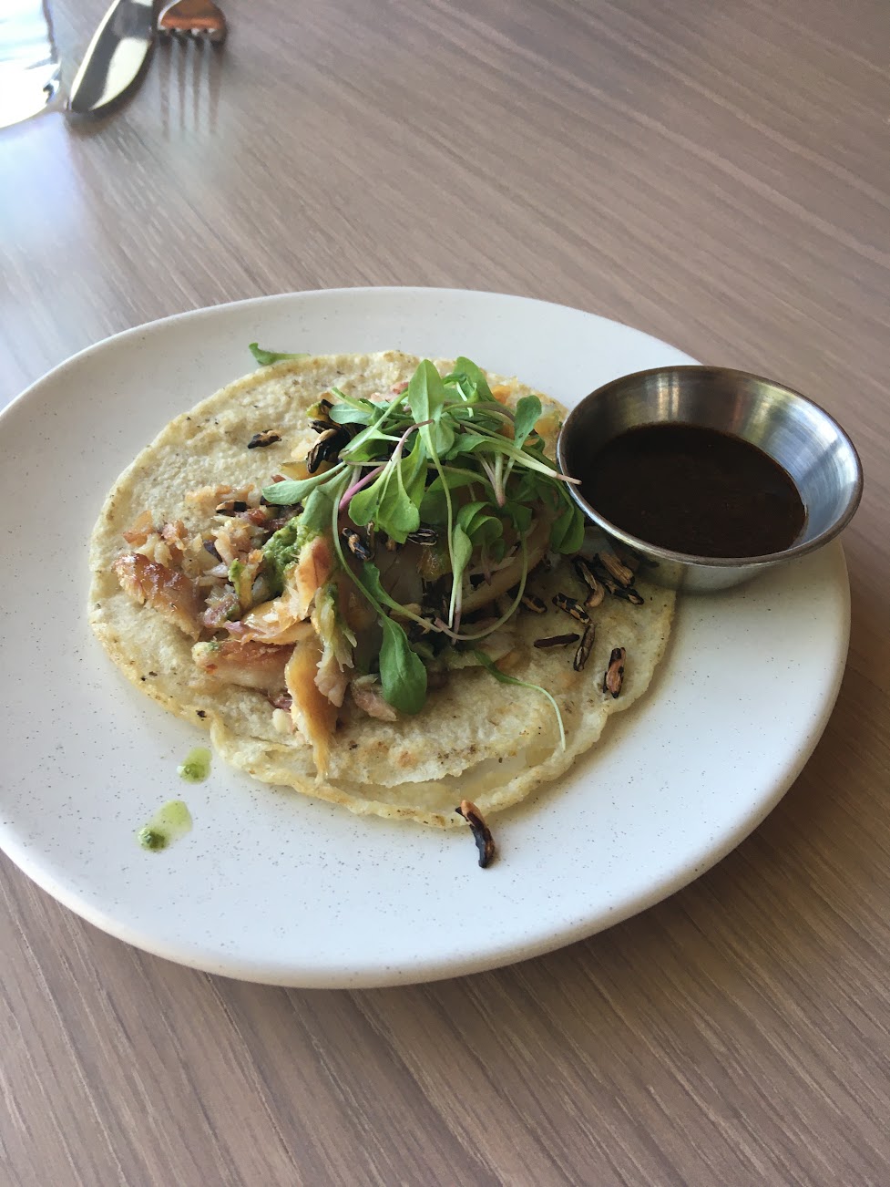 Open faced taco with toppings sits on white plate. Aluminum condiments container with brown sauce sits on plate